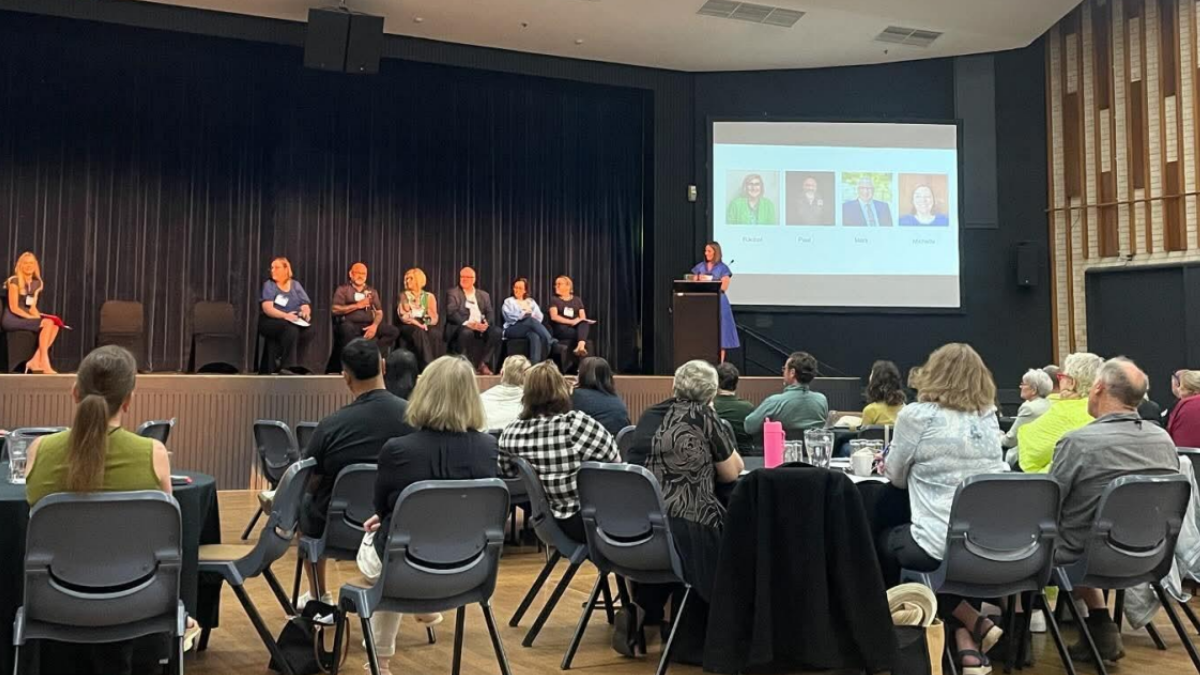 People sitting on chairs at the conference. Some people are on the stage. There is a presentation on a screen.