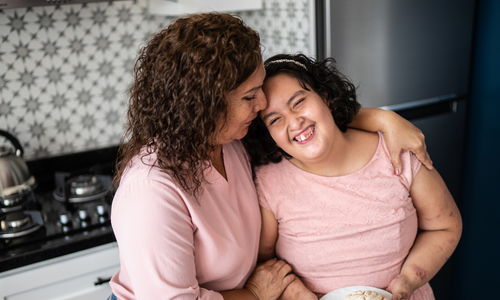 A mother and her daughter with intellectual disability standing in a kitchen. The mother has her arm around her daughter. Her daughter is smiling.