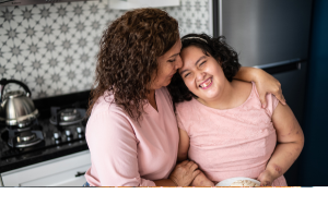 A mother and her daughter with intellectual disability standing in a kitchen. The mother has her arm around her daughter. Her daughter is smiling.
