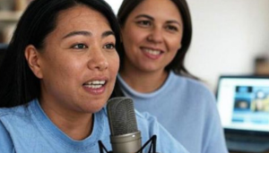 Photo of two people wearing blue tops in a studio. One is talking into a microphone.