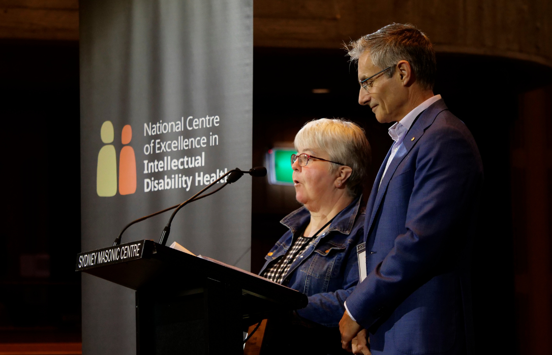 Photo of Julian Trollor and Judy Huett standing on a stage. They are addressing an audience.