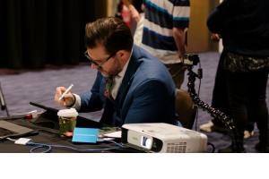 Photo of a man in a suit sitting at a desk. He is writing.