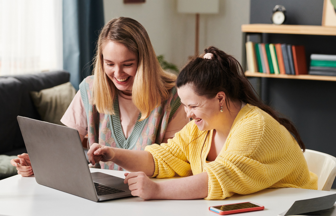 A woman and a girl with intellectual disability at a desk. They are looking at a laptop and smiling.