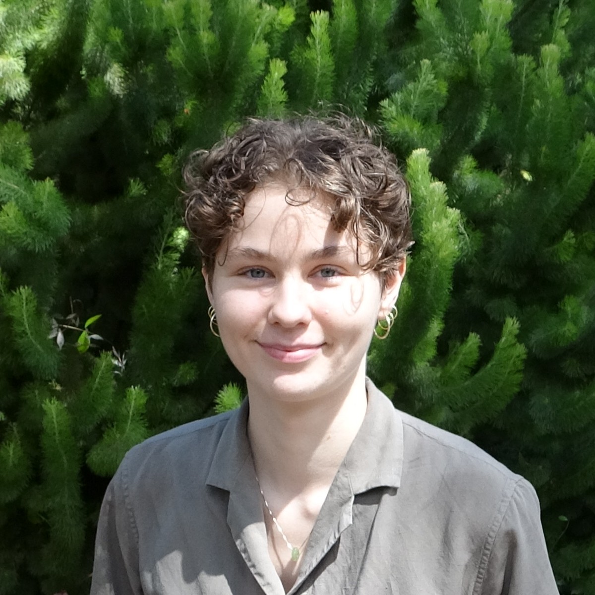 Photo of Annelise Ryan. She is standing in front of green plants and wearing a grey top. She is smiling at the camera.