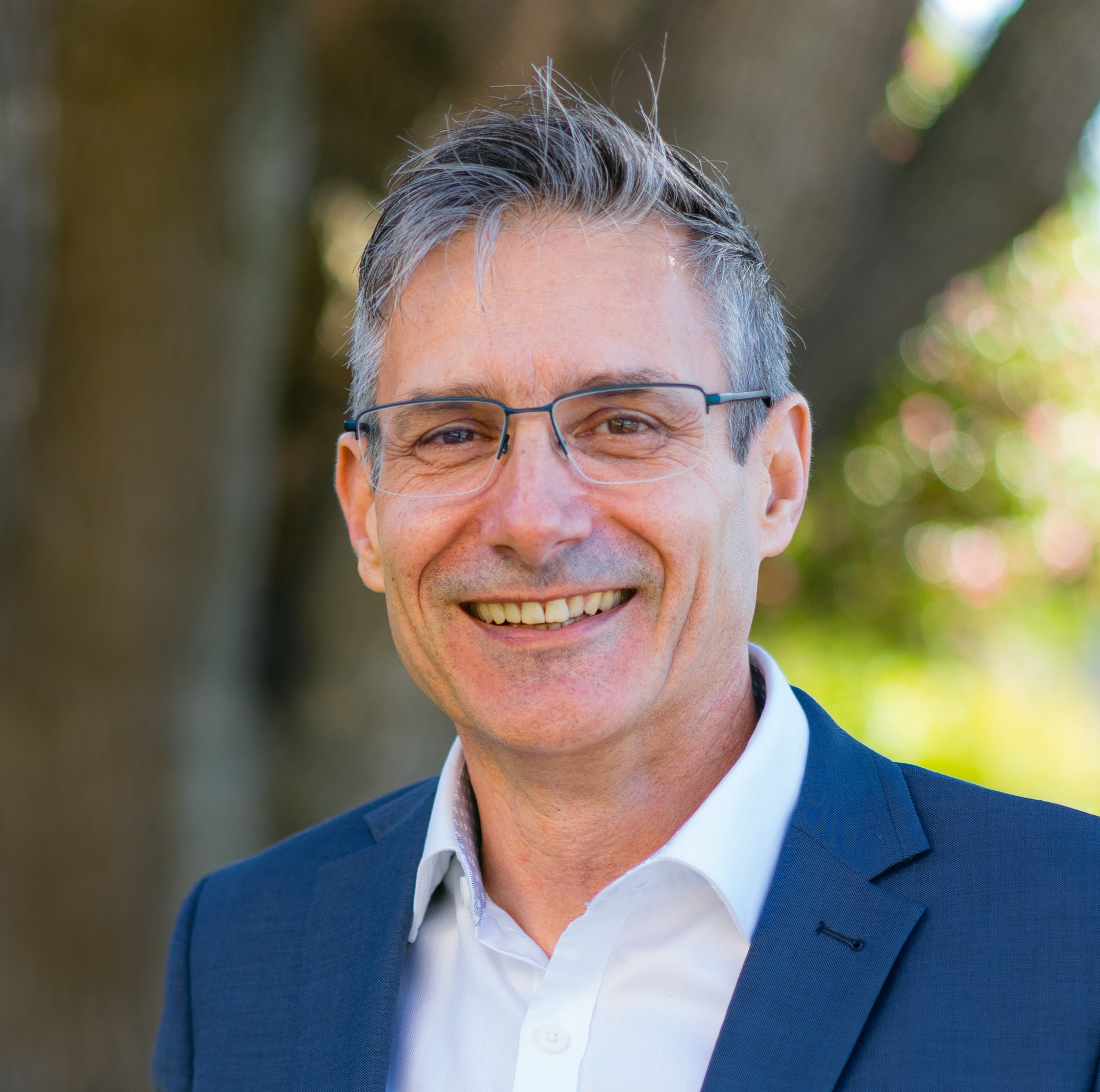 Photo of Julian Trollor. He is standing in front of a tree. He is wearing a blue suit and smiling at the camera.