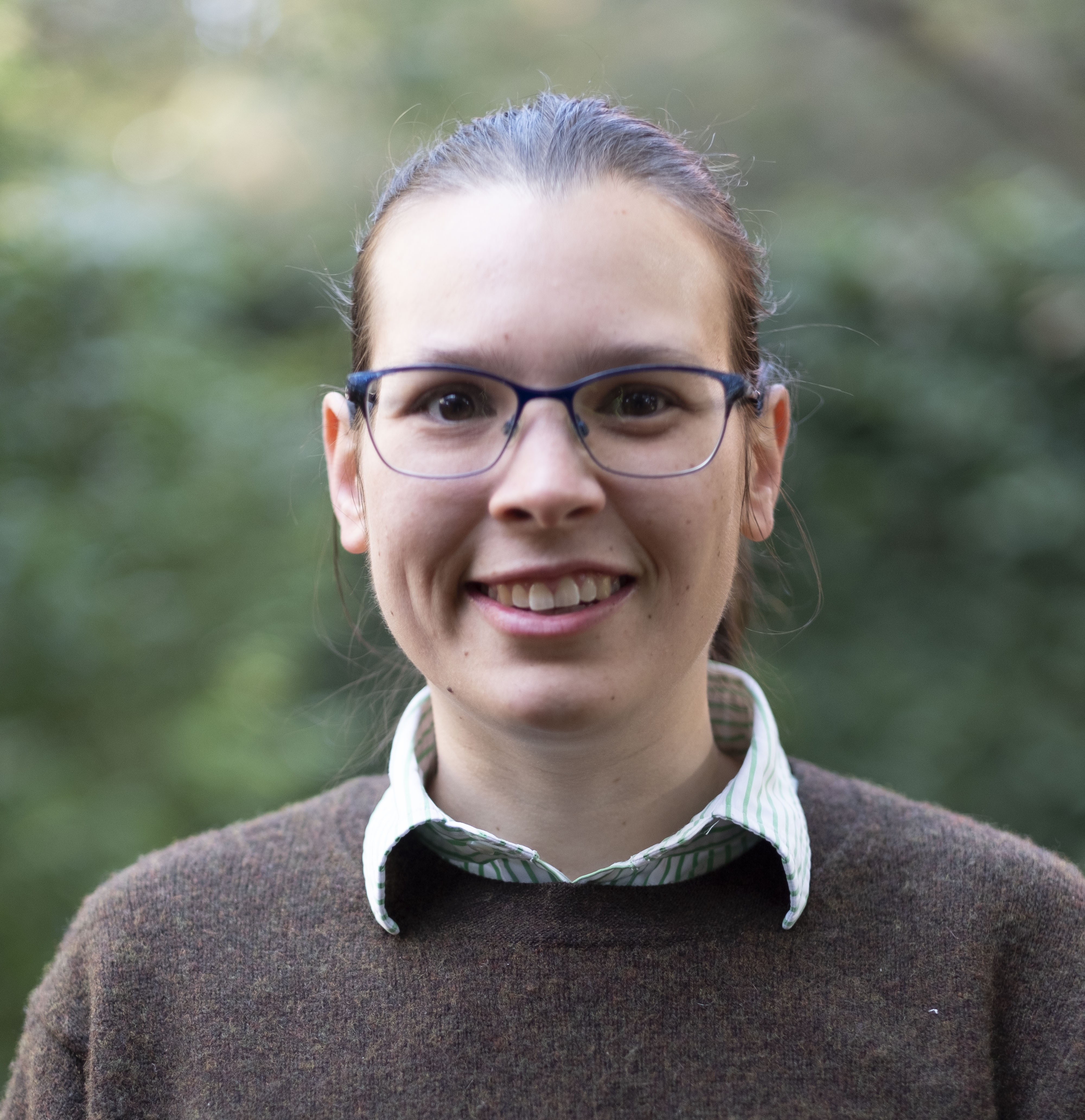 Photo of Tahli Hind. She is standing in front of green plants. She is wearing a brown top and smiling at the camera.
