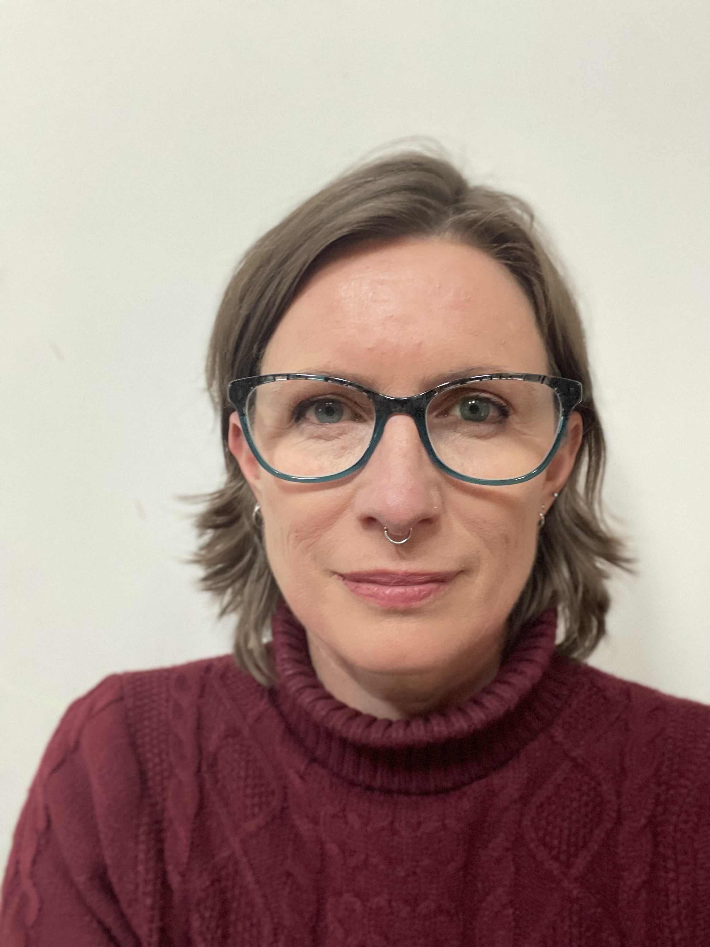 Photo of Danielle Carey. She is wearing a maroon top and glasses. She is smiling at the camera.