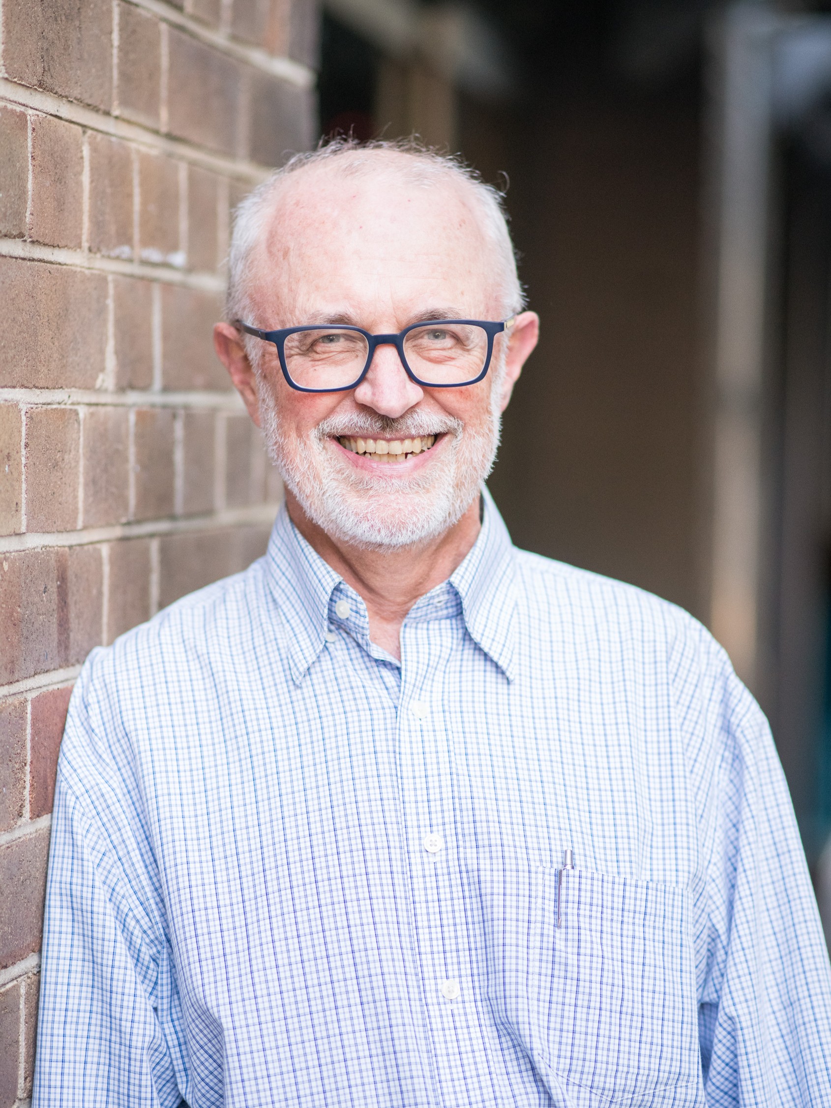 Photo of Jim Simpson. He is wearing a blue top and glasses. He is leaning against a brick wall and smiling at the camera.