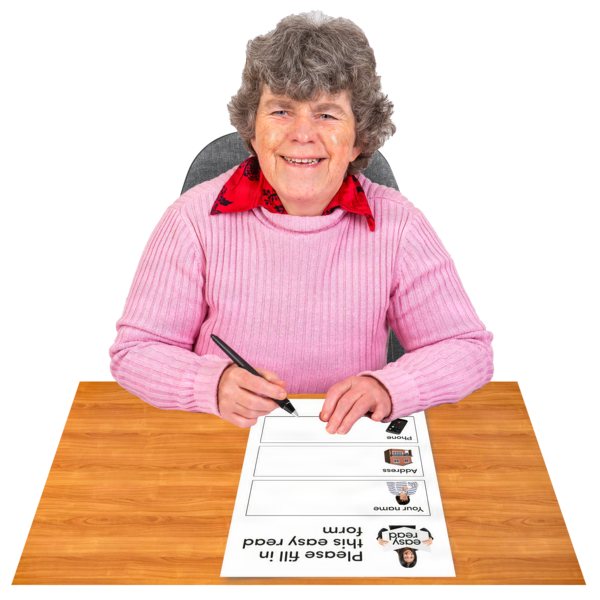 A woman with a pink jumper sitting at a desk. She has grey hair. She is writing on an Easy Read document.