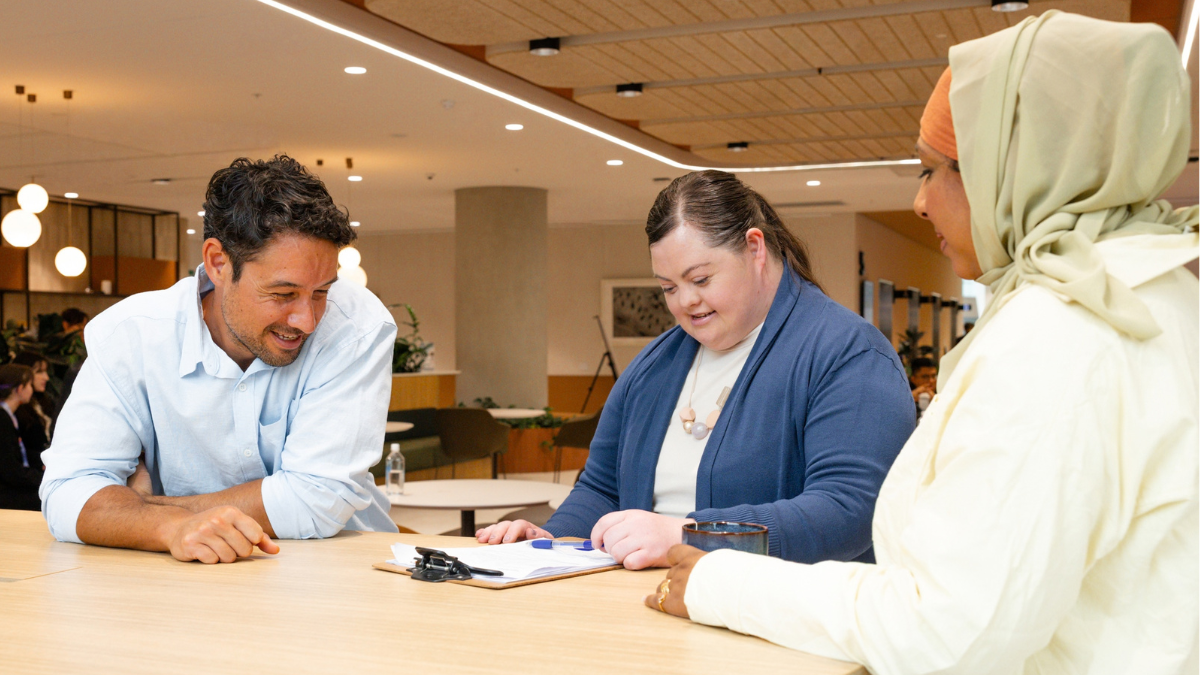 A girl with intellectual disability at a table. She is looking at a clipboard. There is a man on the left. He is leaning on the table and smiling. There is a woman on the right. She is talking to the girl.