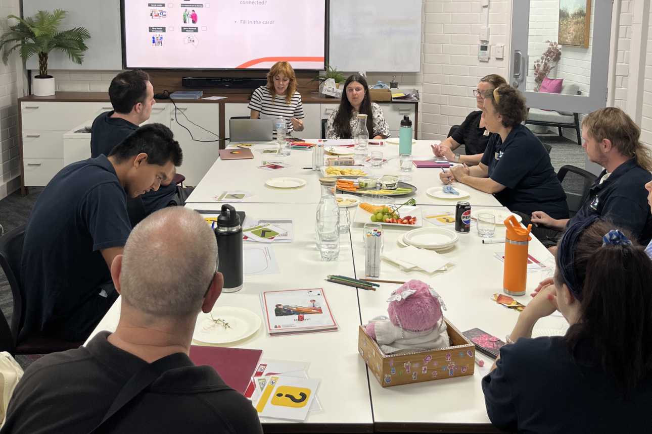 Photo of a group of people sitting around a table. There is a big screen at the front of the table. They are looking at the screen.