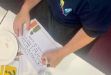 Photo looking down at someone's arms. They are sitting at a desk and filling out a worksheet.