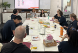 Photo of a group of people sitting around a table. There is a big screen at the front of the table. They are looking at the screen.