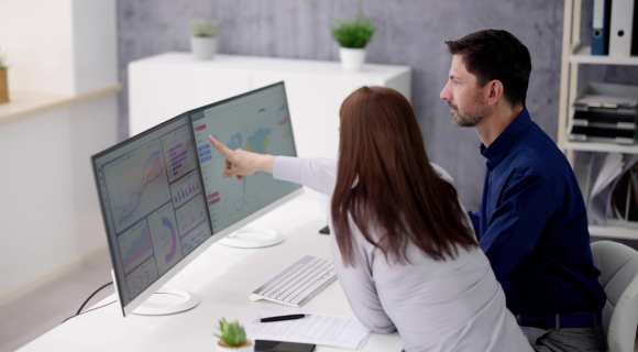 Photo of a man and a woman sitting at a desk looking at data on a computer. The woman is pointing at the computer screen.