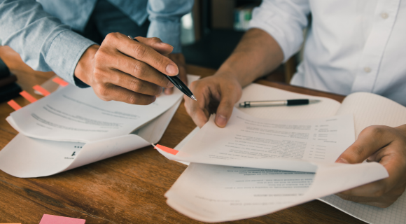 Photo of some official-looking papers on a desk and hands of two people going through them.