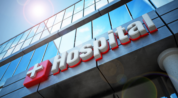 Photo looking up at one side of a hospital. The word Hospital and a cross on the front of the building.