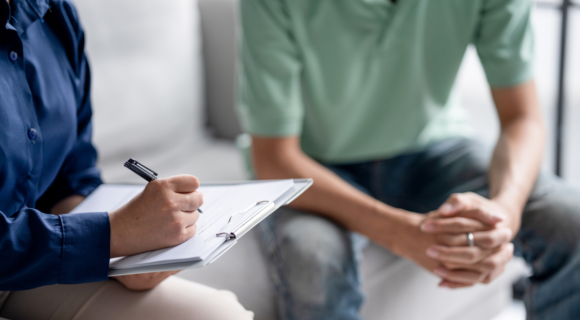 Photo of a psychologist and a client. The psychologist is holding a pen and a clipboard. You cannot see their faces in the photo.