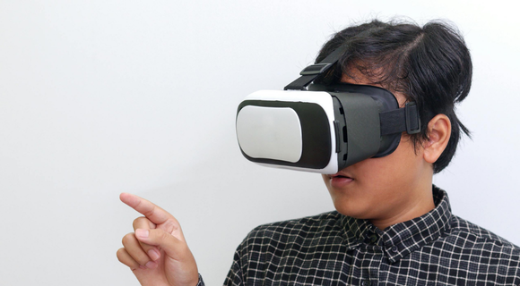 Photo of a man with with a virtual reality headset on. The headset is big. It covers his eyes. He is pointing at something.