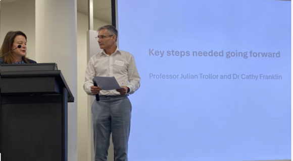 Julian Trollor and Cathy Franklin standing at a podium. A big screen with the words "Key steps needed going forward" is behind them.