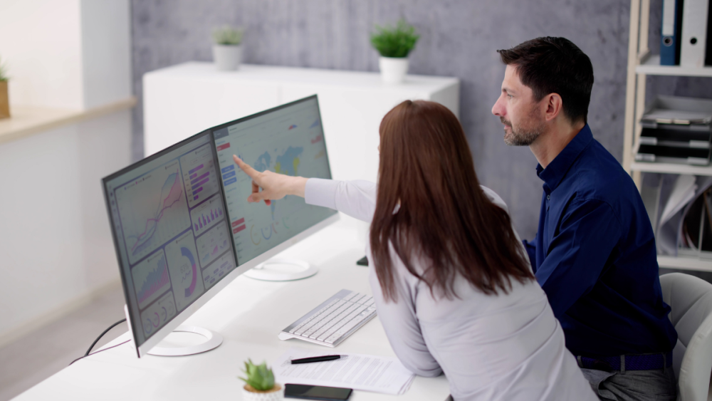 Photo of a man and a woman sitting at a desk looking at data on a computer. The woman is pointing at the computer screen.