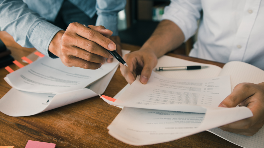 Photo of some official-looking papers on a desk and hands of two people going through them.