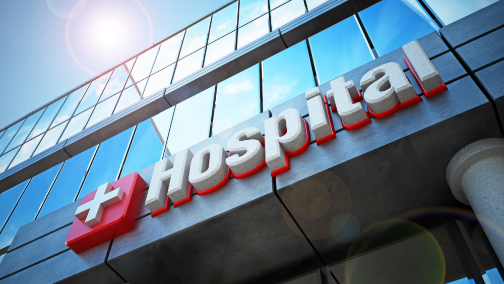 Photo looking up at one side of a hospital. The word Hospital and a cross on the front of the building.