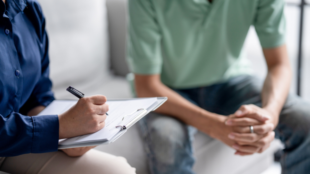 Photo of a psychologist and a client. The psychologist is holding a pen and a clipboard. You cannot see their faces in the photo.