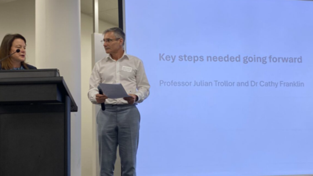 Julian Trollor and Cathy Franklin standing at a podium. A big screen with the words "Key steps needed going forward" is behind them.