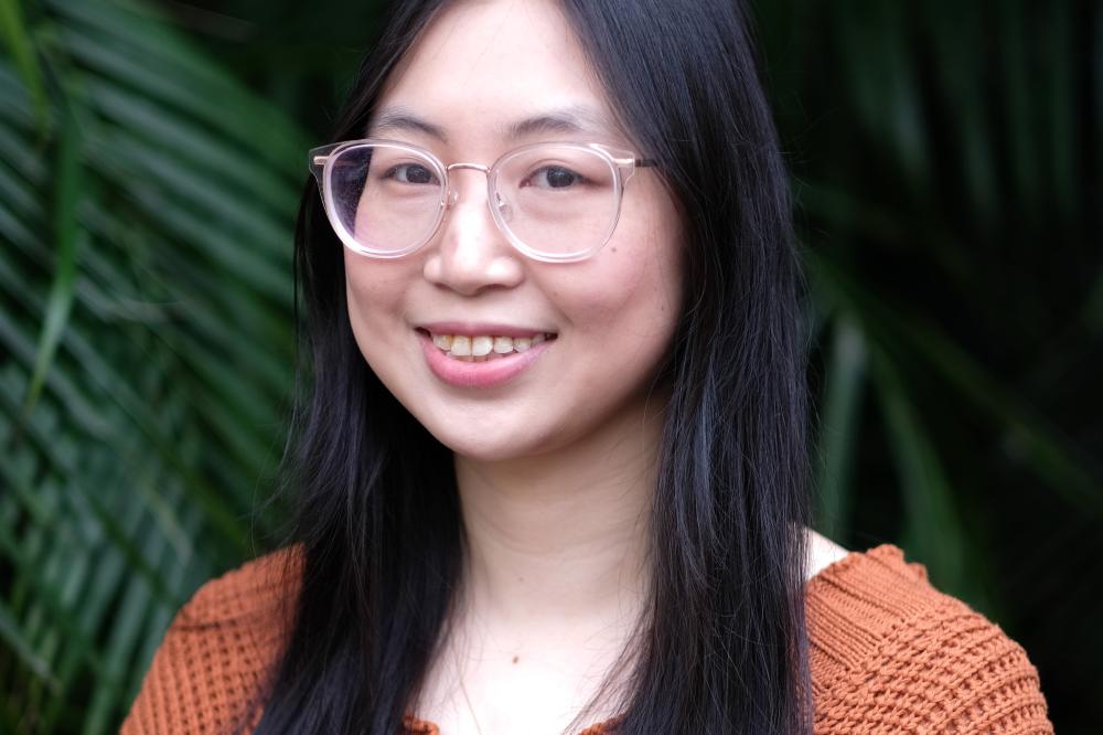A photo of Jenna Zhao. The photo is of Jenna's shoulders and head. She is wearing glasses and smiling at the camera.