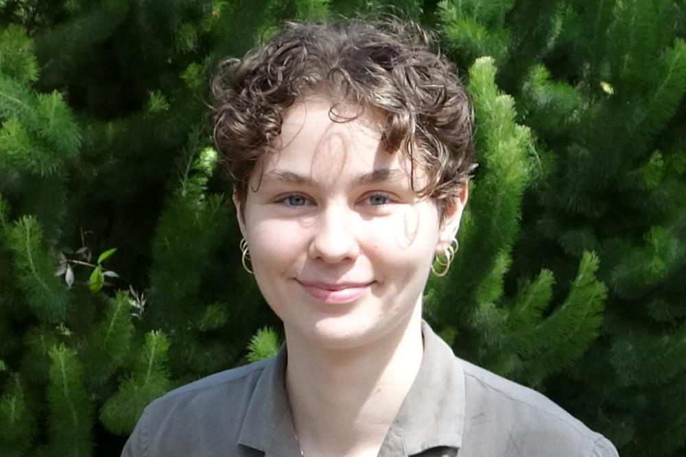 Photo of Annelise Ryan. She is standing in front of green plants and wearing a grey top. She is smiling at the camera.