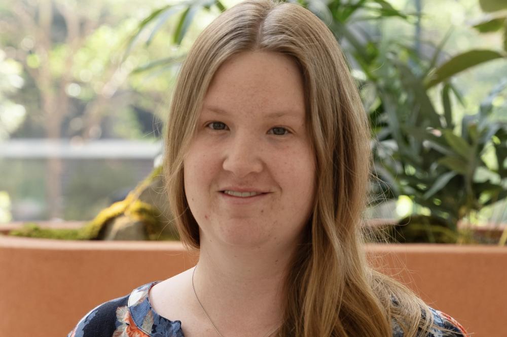 Photo of Madison Booth. She is sitting in front of a green plant and smiling at the camera.