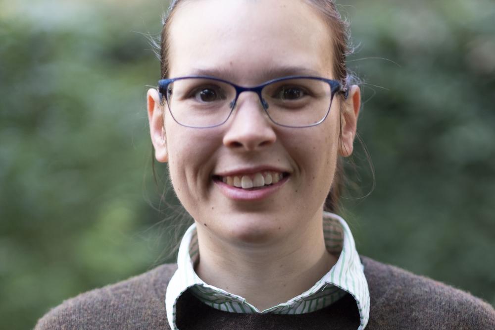 Photo of Tahli Hind. She is standing in front of green plants. She is wearing a brown top and smiling at the camera.