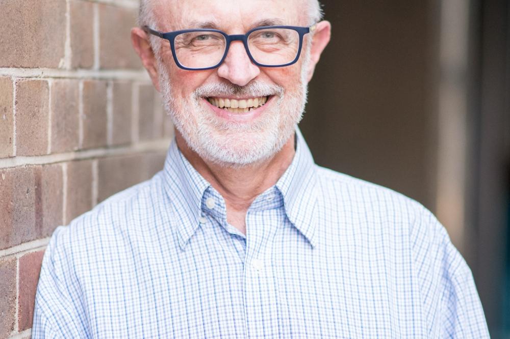 Photo of Jim Simpson. He is wearing a blue top and glasses. He is leaning against a brick wall and smiling at the camera.